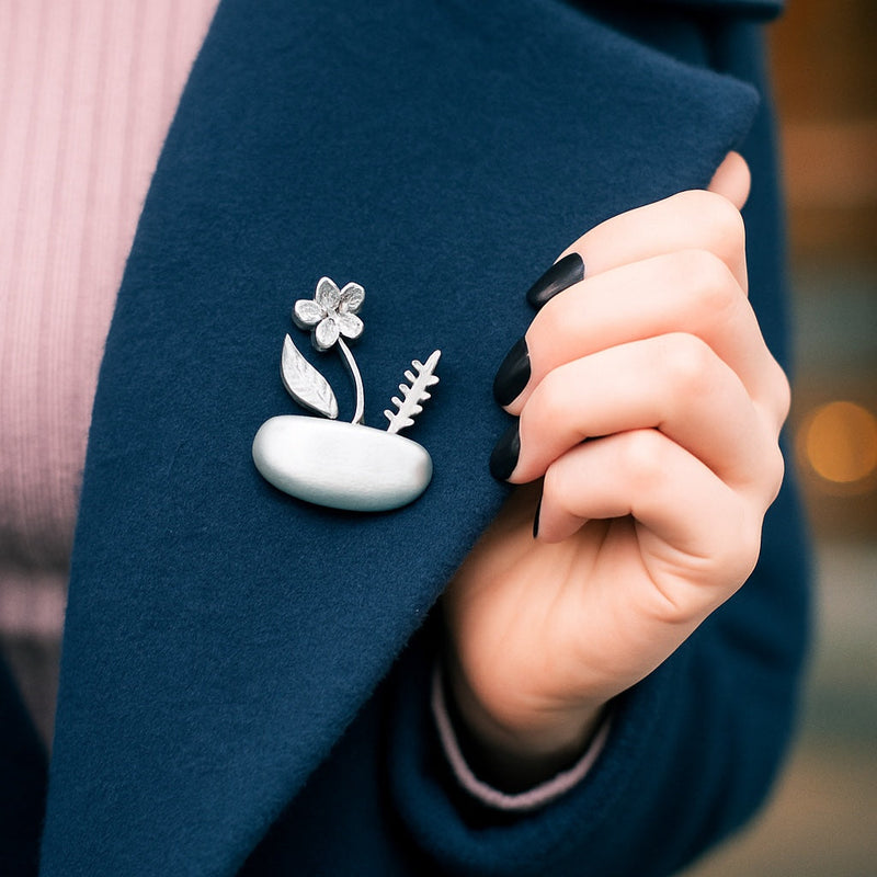 Close-up of a woman in a navy blue coat, with a silver brooch on the lapel. The brooch features a minimalist botanical design: a smooth oval base with a delicate flower, a leaf, and a fern-like sprig rising from it