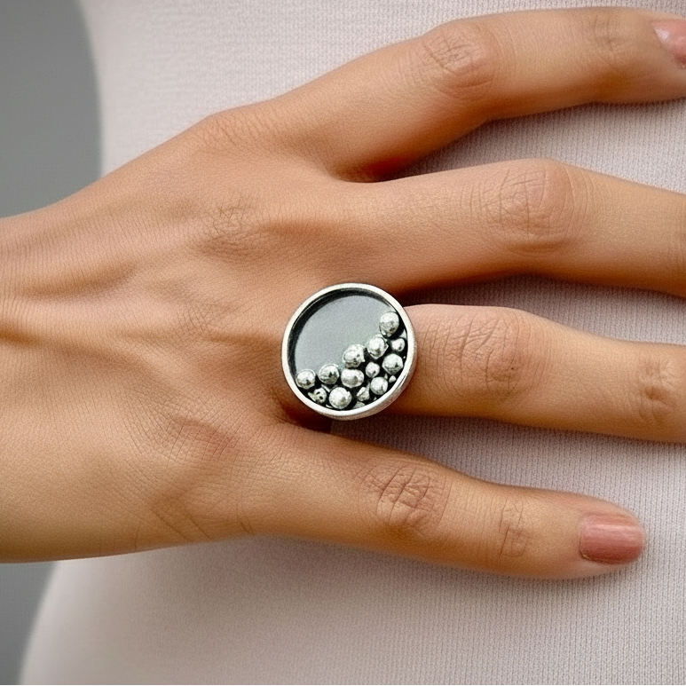 Close-up of a hand wearing a round silver statement ring, featuring a smooth circular face with raised clustered bead details on one side, against a light neutral fabric background