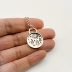 Silver necklace with a round pendant which has trees and sum impressions held between fingers on a white background