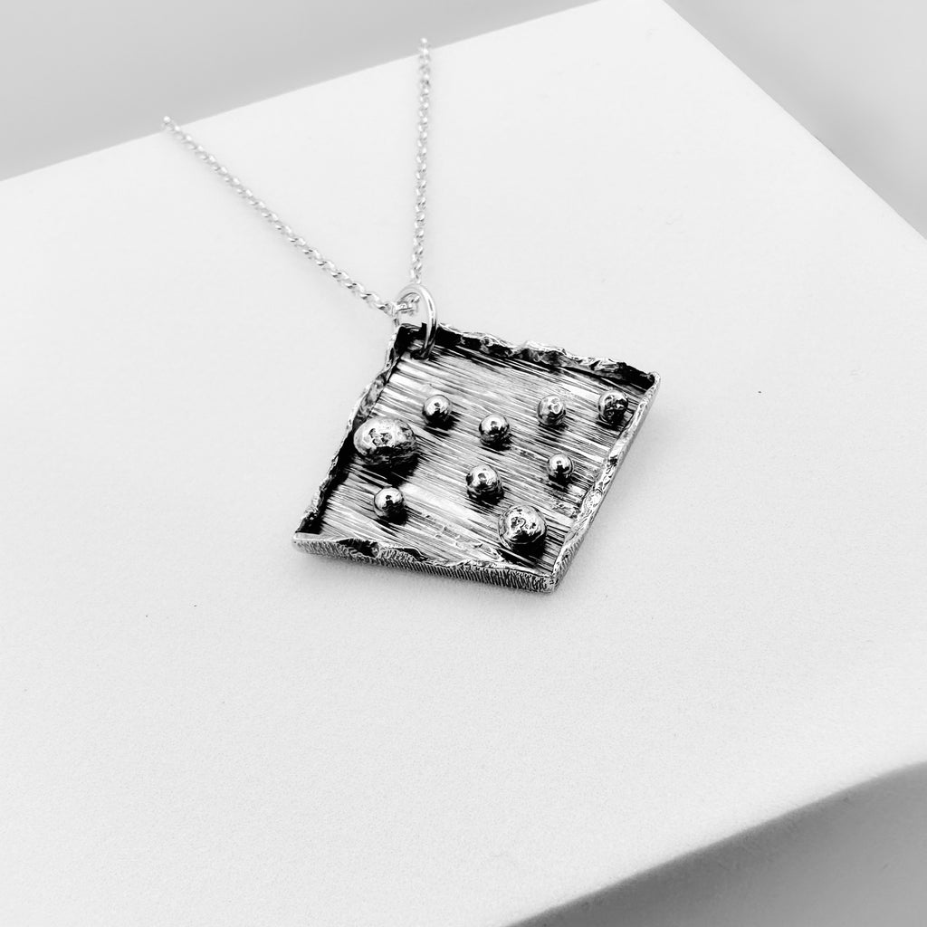 Handmade in Nottingham by Bottlebee a sterling silver necklace with a textured square pendant with silver balls on a white surface
