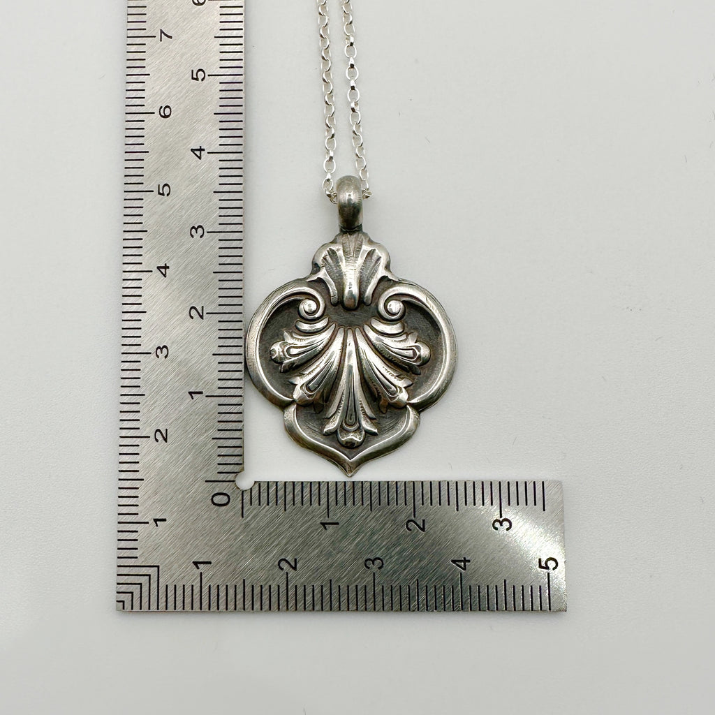Silver necklace with an ornate pendant next to a ruler on a white background