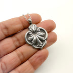 Silver bespoke sustainable ornate jewellery 925 fleur-de-lys silver pendant held between fingers on a white background