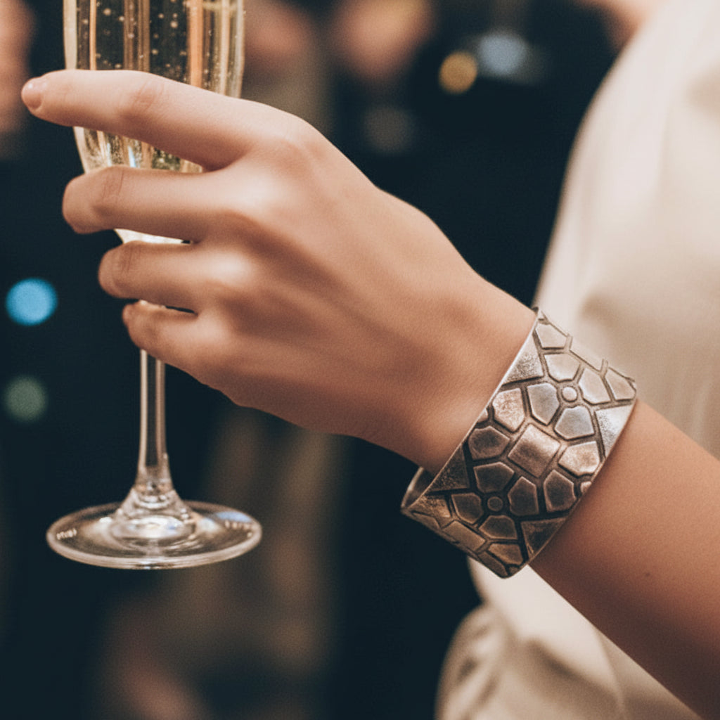 Womans hand wearing a handmade and oxidised geometric patterned wide cuff bangle holding a glass of champagne with a blurred background. Jewellery is handmade and sustainable using recycled sterling silver by a small one woman business in Nottingham