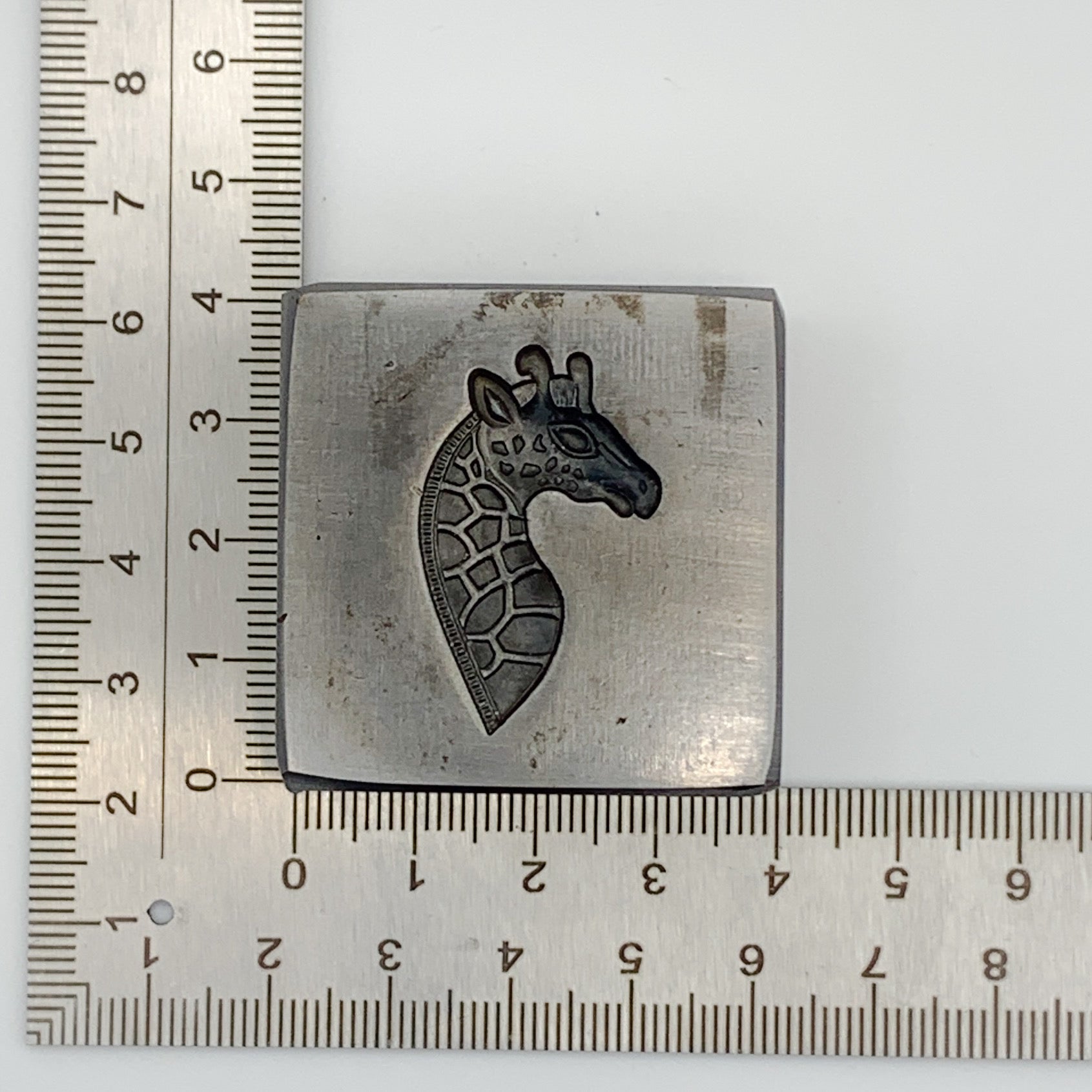 Metal giraffe stamp with giraffe design next to a ruler on a white background the silver will be pressed into the impression die to make the pendant