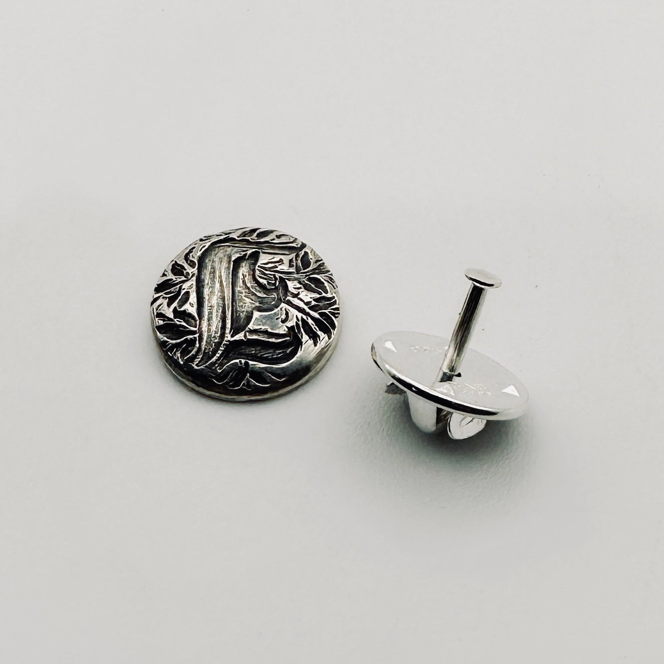 Silver decorative lapel pin with intricate personalised design on a light gray background