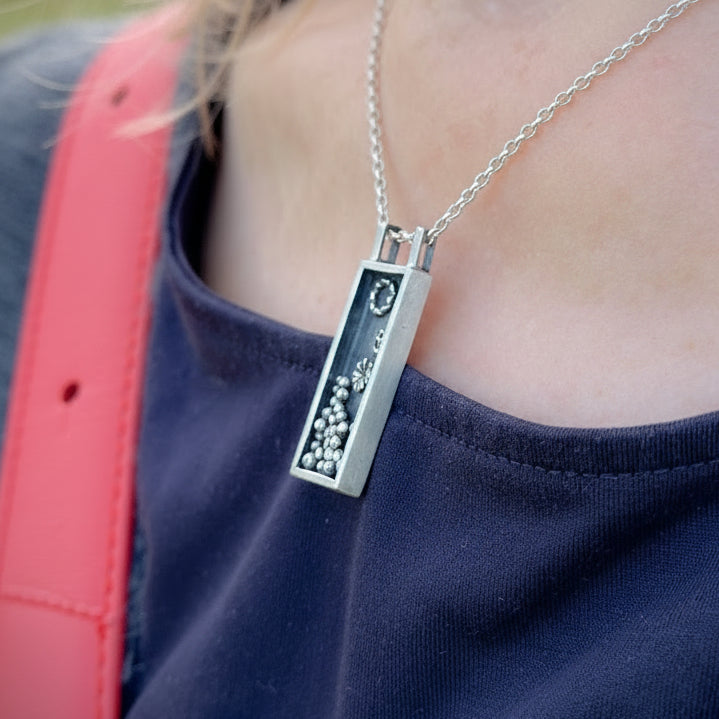 Person wearing a silver necklace with a rectangular pendant outdoors. Necklace make by Bottlebee whose studio is in Nottingham