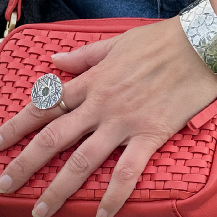 Close-up of a womans hand wearing a wide, textured silver cuff bracelet and a large, round silver kinetic ring, resting on a bright red woven leather handbag.