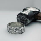 Silver hatch textured 6mm wide wedding ring next to a metal hammer used to create the texture on a light grey background