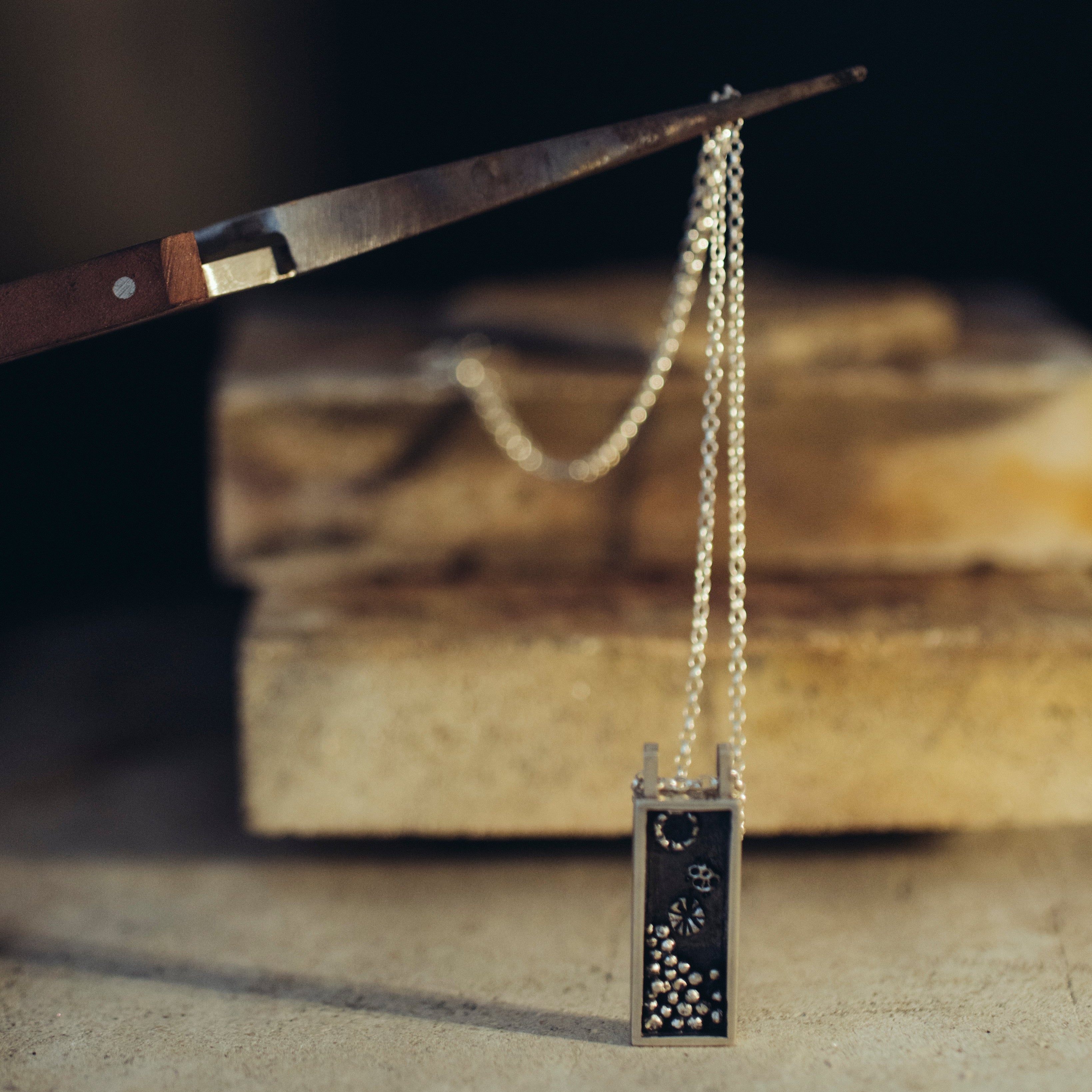 Box necklace with an oxidised pendant with granules on a soldering surface with a blurred background being held by tweezers