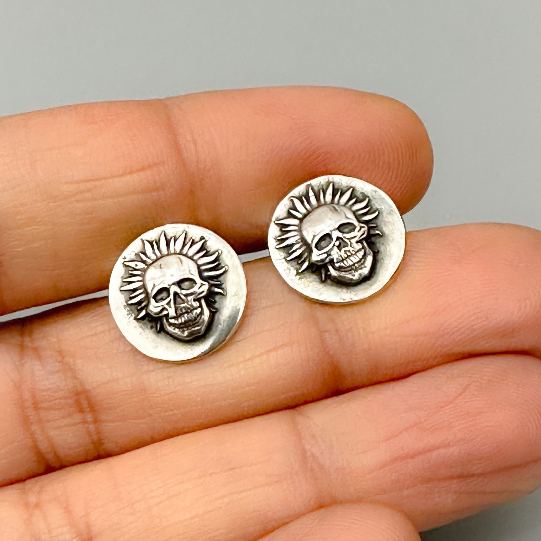 Funky sustainable original sterling silver skull earrings held between fingers against a neutral background handmade by small business called Bottlebee in Nottingham