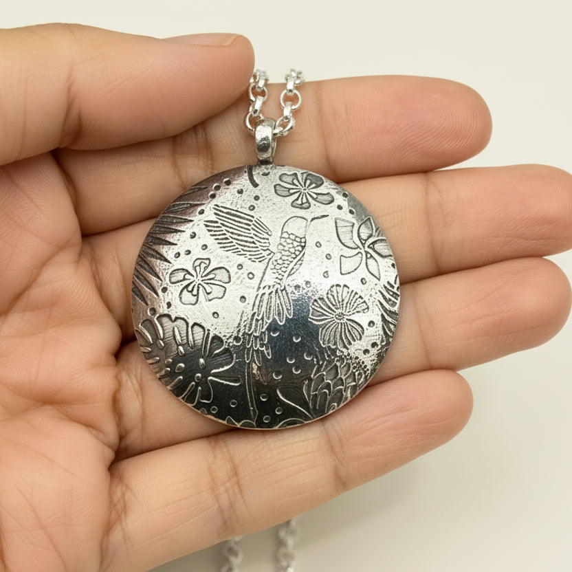 Large round silver pendant with intricate designs of hummingbird, leaves and flowers held in a hand sustainable jewellery handmade in nottingham by bottlebee