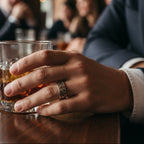 Man holding a glass of whiskey with a blurred bar setting in the background. Wearing a silver wedding band made by Bottlebee in Nottingham sustainable jewellery