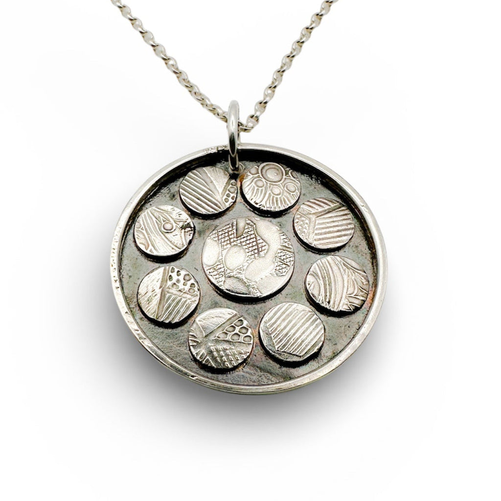 Silver necklace with a round pendant featuring embossed designs on a white background handmade sustainable jewellery by Bottlebee in nottingham