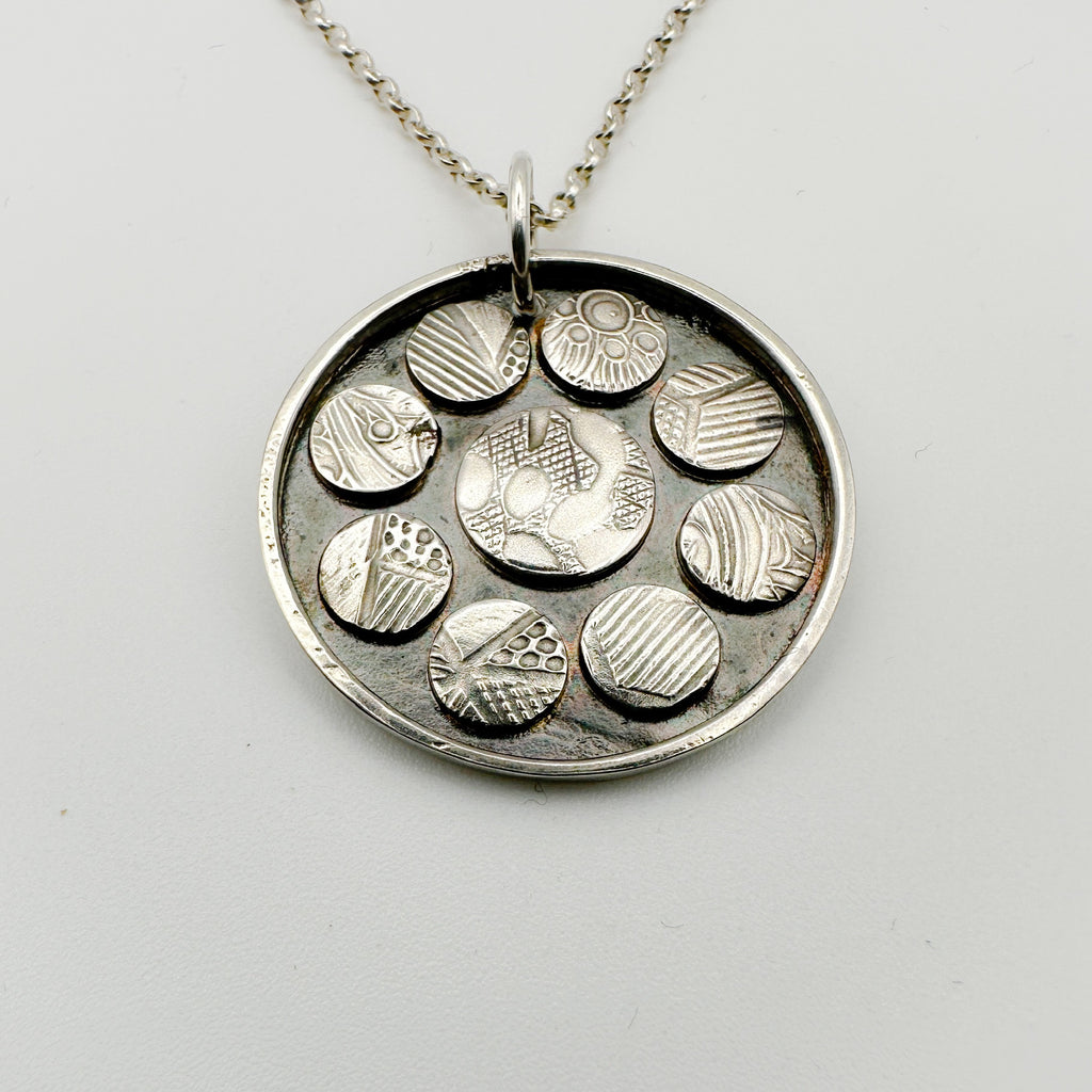 Silver necklace with a round pendant featuring embossed circular designs on a light grey background