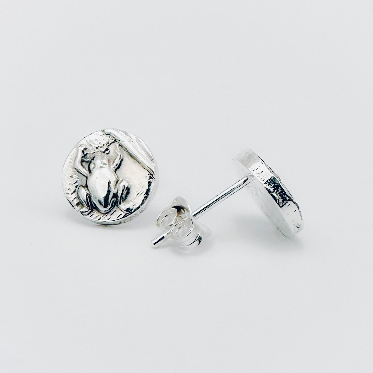 Silver round earrings with a frog impression on a light grey background