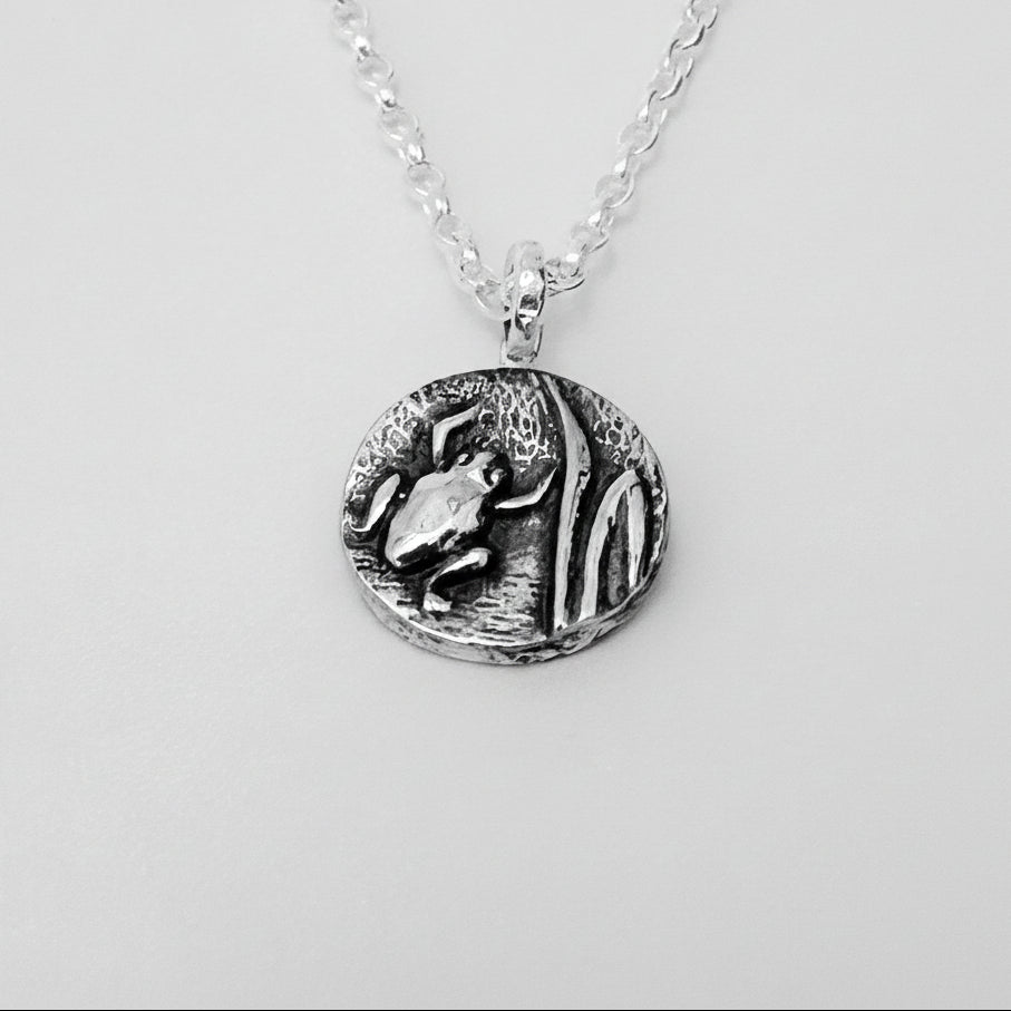 A sterling silver disc pendant with a frog and leaf protruding out of the silver disc and the pendant is on a silver belcher chain, jewellery is handmade by Bottlebee in Nottingham