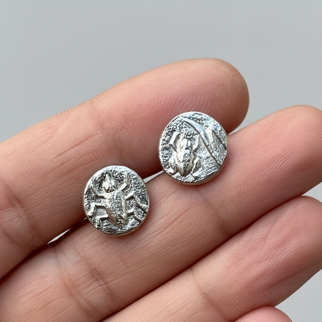 a pair of handmade silver stud earrings resting on fingers, one earring. has a frog the other one is a beetle, both images are raised on the disc