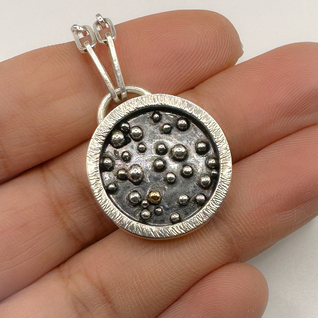 Silver pendant with a granulated 18ct gold ball and sterling silver balls which has been oxidised held between fingers on a white background
