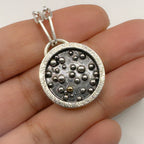 Silver pendant with a granulated 18ct gold ball and sterling silver balls which has been oxidised held between fingers on a white background
