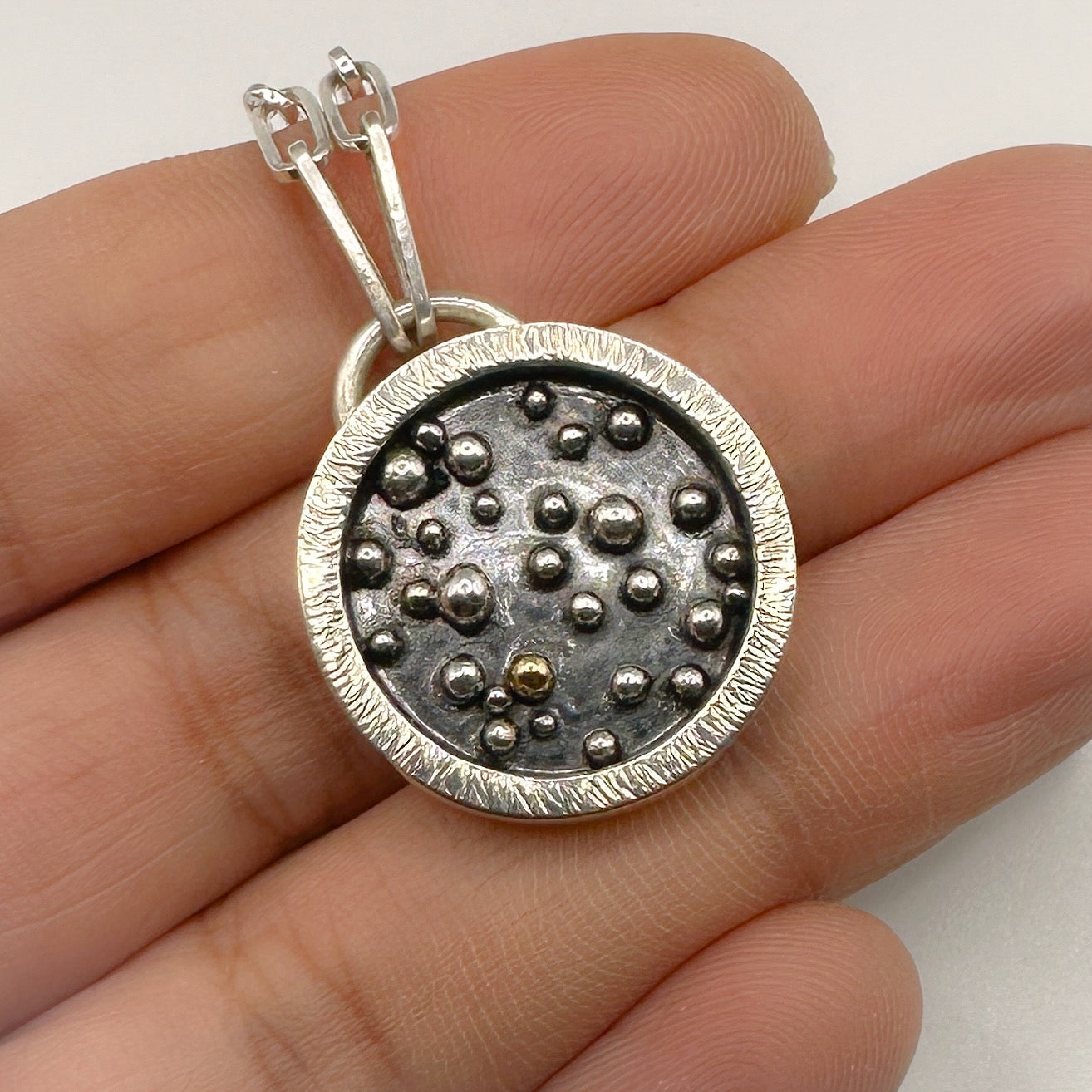 Silver pendant with a granulated 18ct gold ball and sterling silver balls which has been oxidised held between fingers on a white background