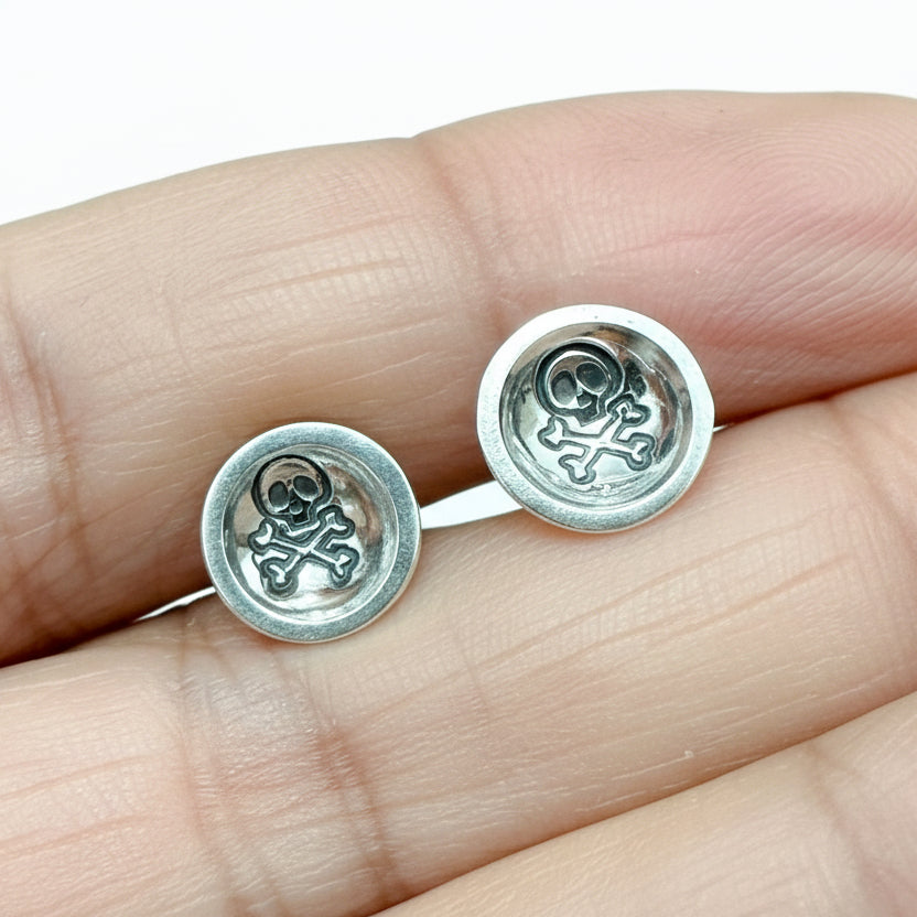 Silver skull and crossbones earrings held between fingers against a white background handmade by a small business called Bottlebee who makes sustainable jewellery in Nottingham