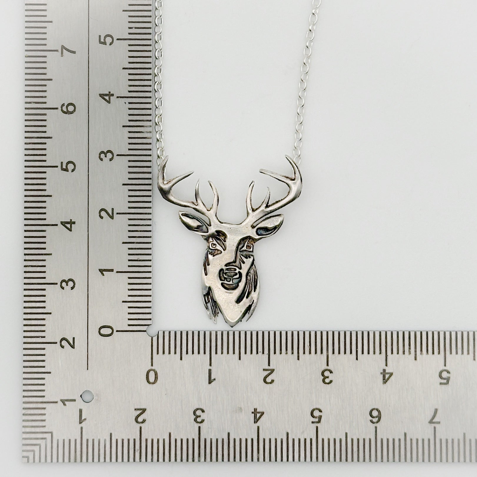 Silver deer head necklace on a white background with a ruler for scale, handmade by Bottlebee in Nottingham