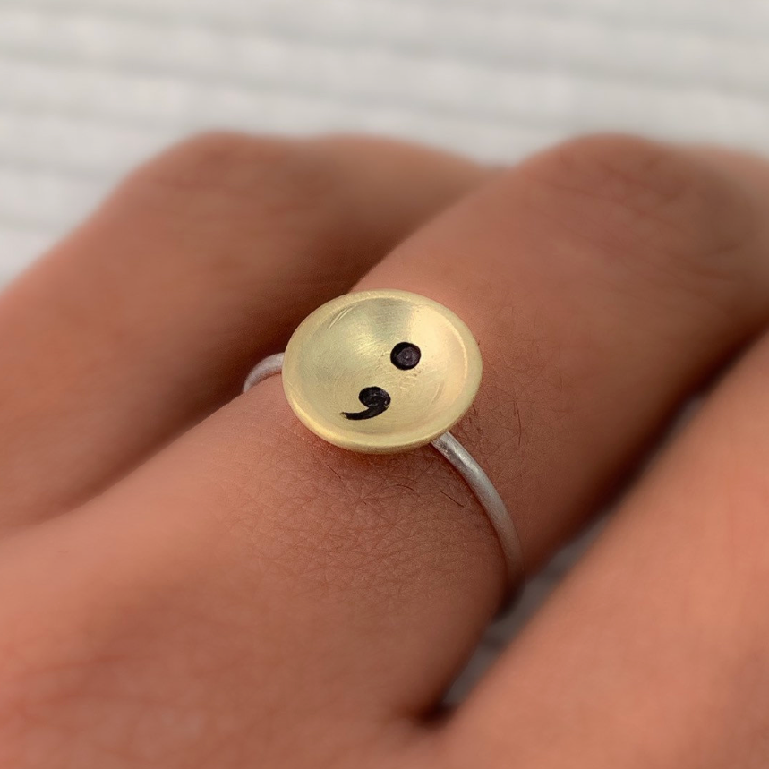 Gold ring with a semi colon design on a person's finger against a blurred background handmade in nottingham