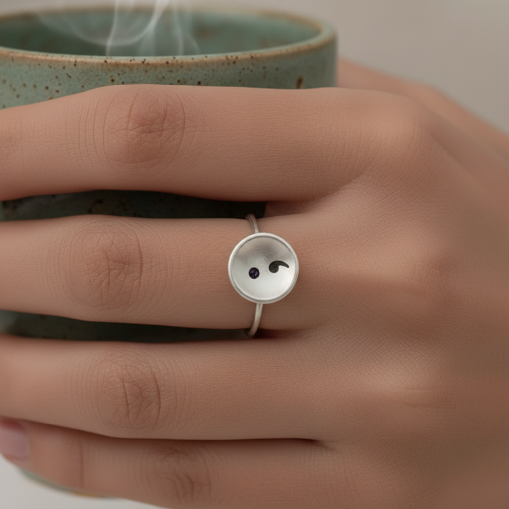 Hand holding a steaming mug with a button-shaped semi colon ring on a neutral background
