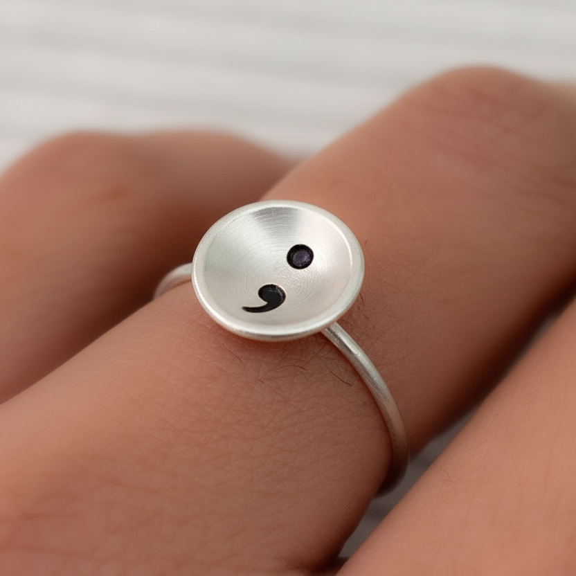 Silver ring with a semi colon design on a finger against a neutral background