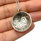 Silver pendant with intricate oxidised floral design held between fingers on a neutral background