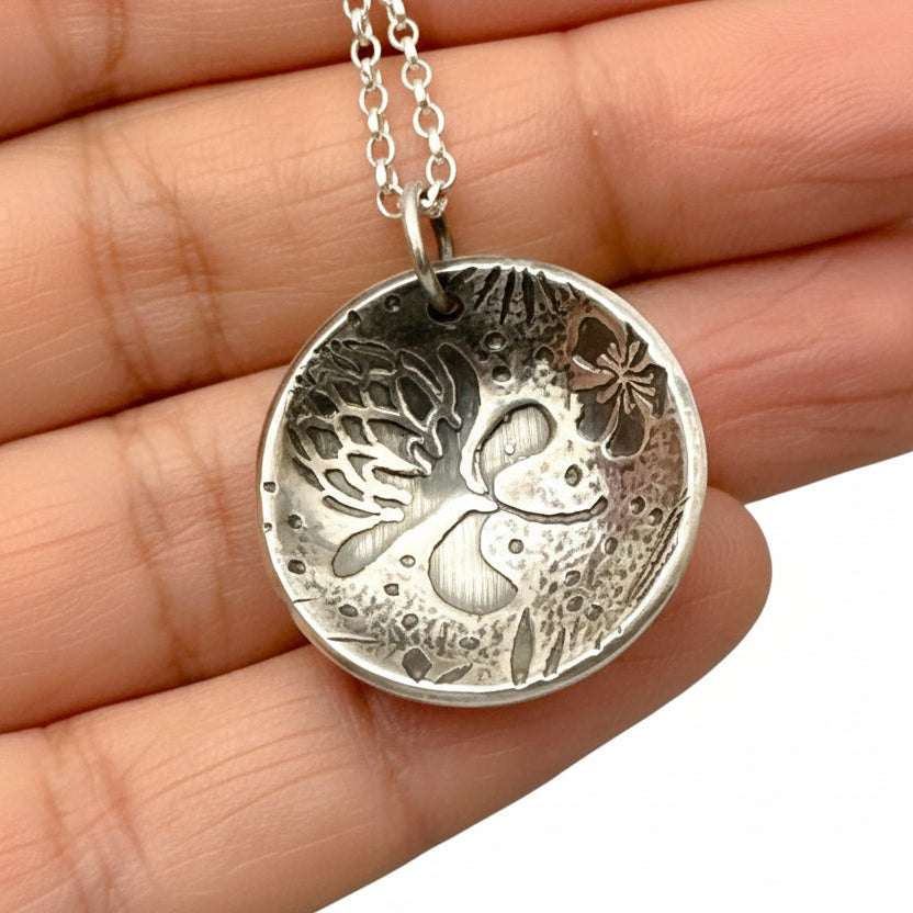 Silver pendant with intricate oxidised floral design held between fingers on a neutral background
