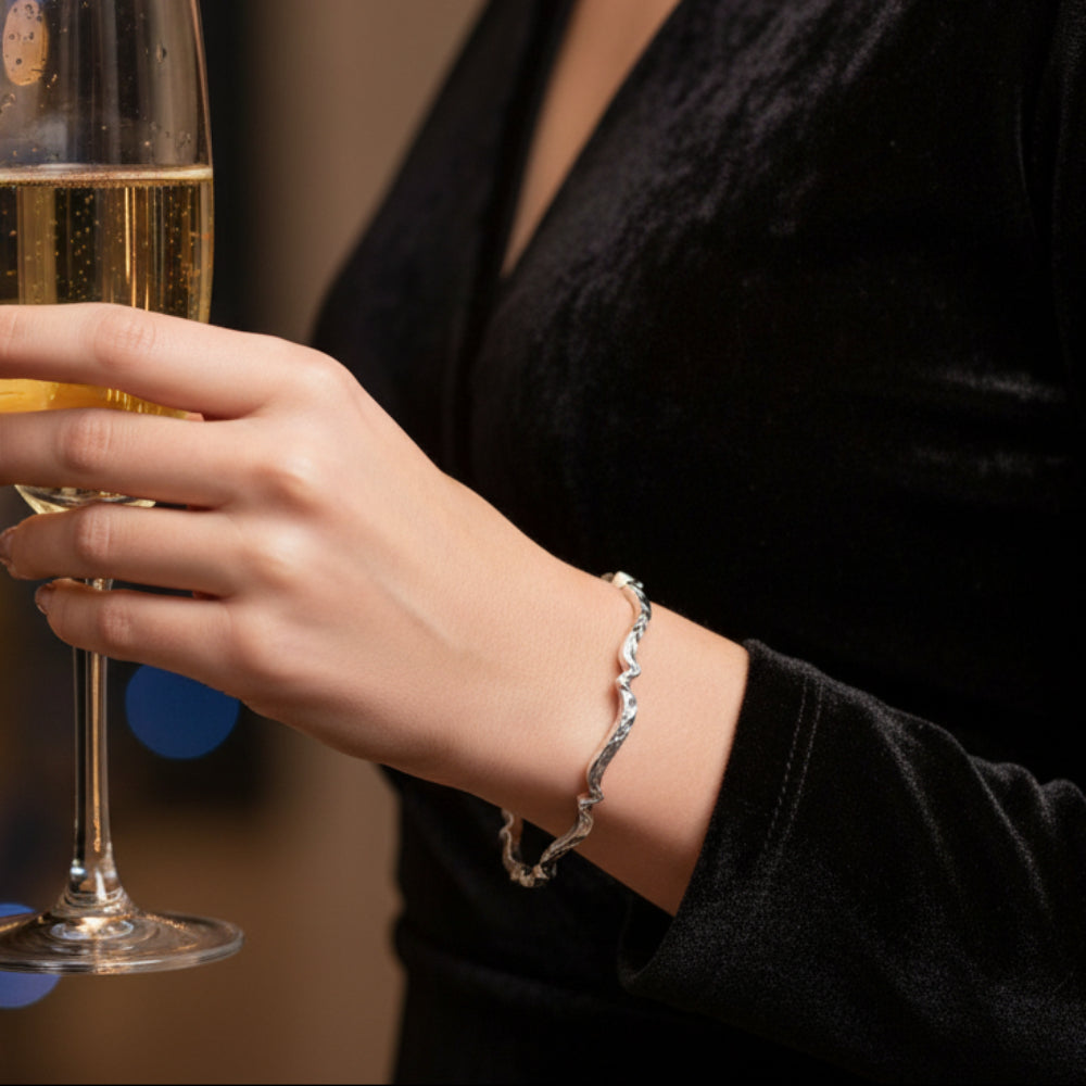 A woman wearing a black top holding a glass of champagne, wearing a handmade luxurious sterling silver wavy bangle with a hammered and oxidised texture