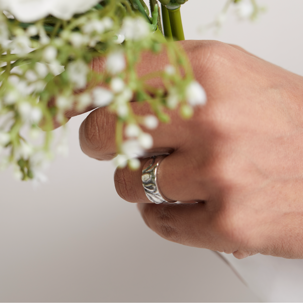 Hand holding a bouquet of white flowers with a bespoke handmade silver wedding ring on a light background