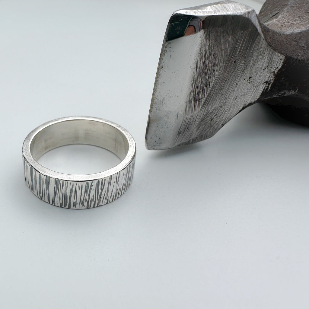 lined unique silver wedding ring band for a man or woman or unisex with textured surface next to a metal hammer on a light grey background handmade sustainable bespoke jewellery nottingham