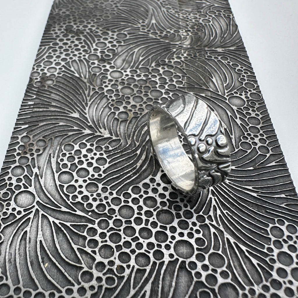 unusual bespoke ocean waves patterned silver wedding ring band for a man or woman or unisex resting on a pattern plate on a light grey background handmade sustainable jewellery Nottingham