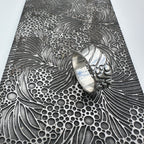 unusual bespoke ocean waves patterned silver wedding ring band for a man or woman or unisex resting on a pattern plate on a light grey background handmade sustainable jewellery Nottingham