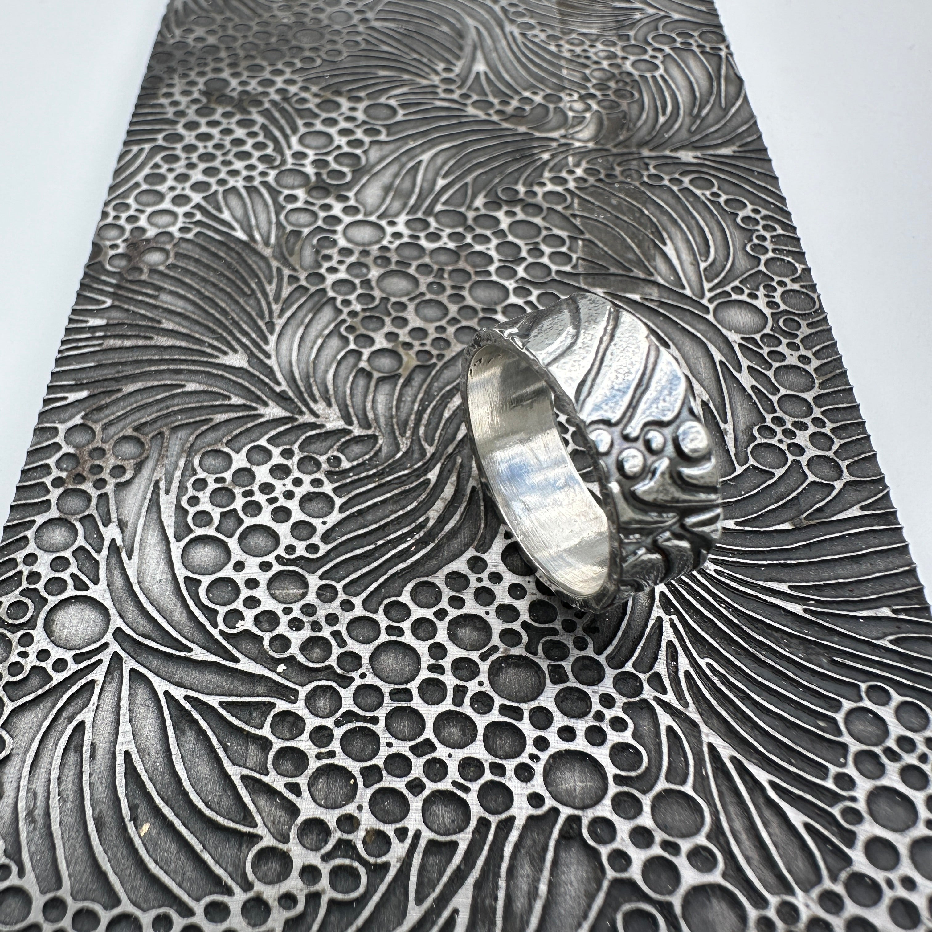 unusual bespoke ocean waves patterned silver wedding ring band for a man or woman or unisex resting on a pattern plate on a light grey background handmade sustainable jewellery Nottingham