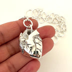 A hand holding silver jewellery which is an anatomical heart pendant with handmade link chain