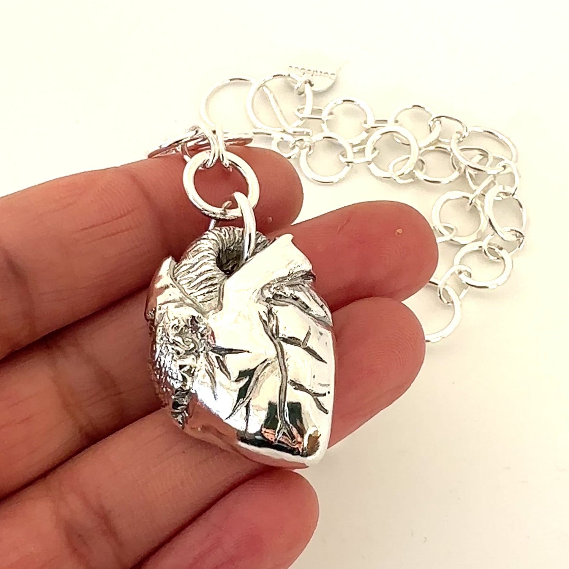 A hand holding silver jewellery which is an anatomical heart pendant with handmade link chain