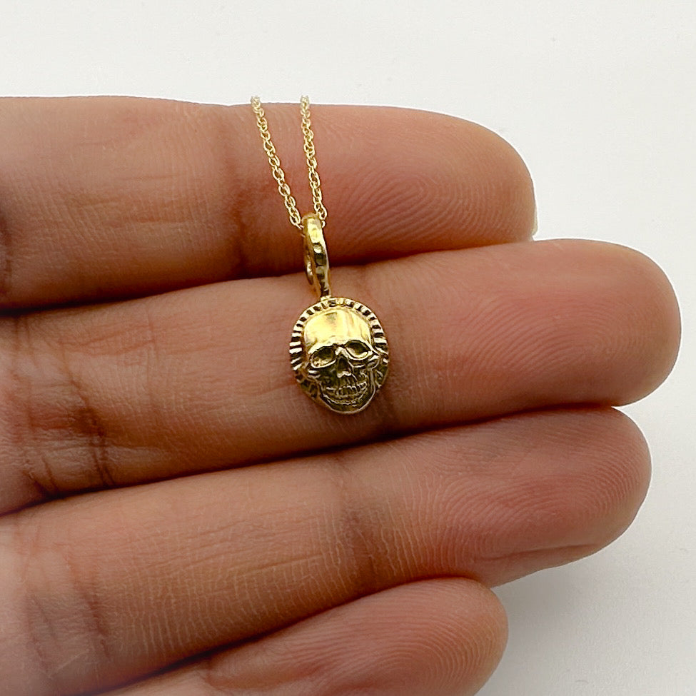 Gold skull pendant necklace held between fingers on a white background