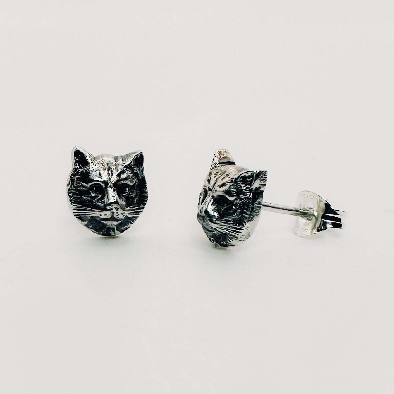 A front facing and side facing pair of silver cat face earrings with a butterfly back handmade in nottingham by Bottlebee